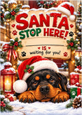 A festive Christmas sign reading 'SANTA STOP HERE! IS waiting for you!' with a Rottweiler dog wearing a Santa hat in the snow below, surrounded by ornaments, lanterns, wrapped gifts, and holiday decorations.