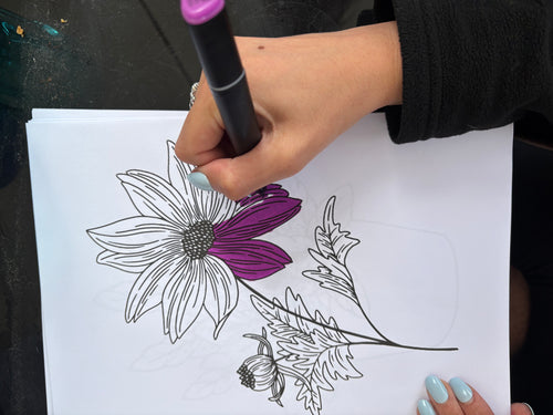 Person coloring a floral design with a purple marker on white paper.