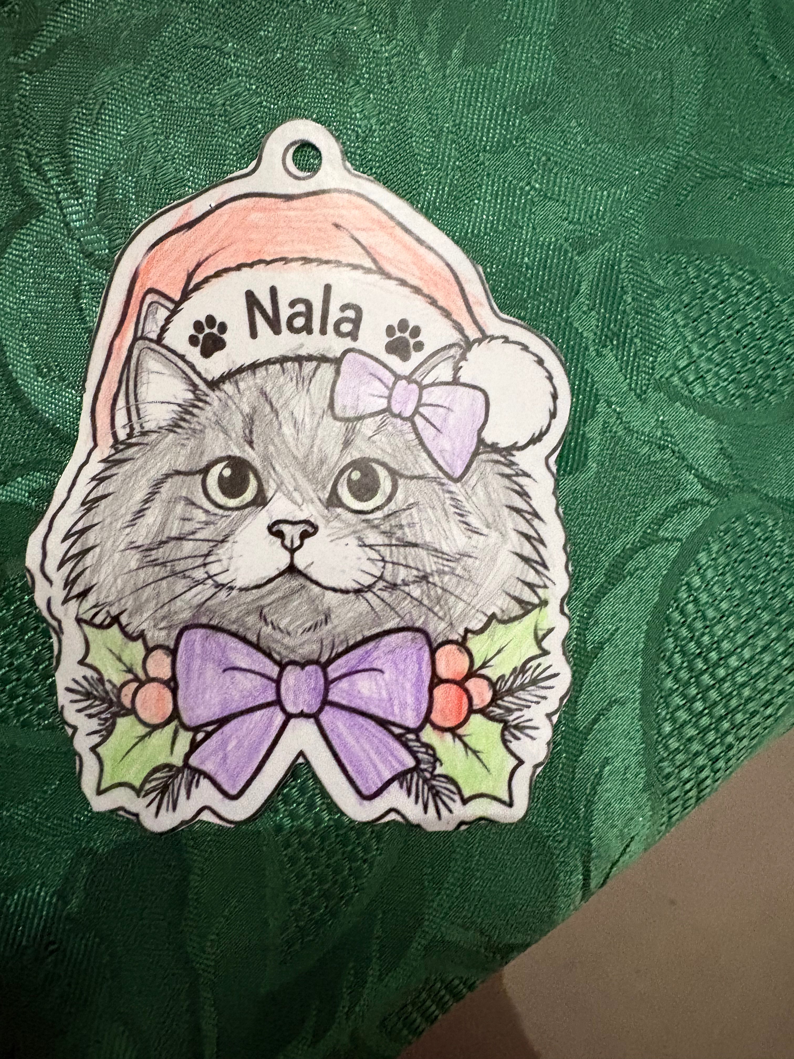 A   cat Christmas tree decoration featuring a hand-drawn gray tabby cat wearing a pink Santa hat and a purple bow, with the name 'Nala' written on the hat, displayed on a textured green fabric background.