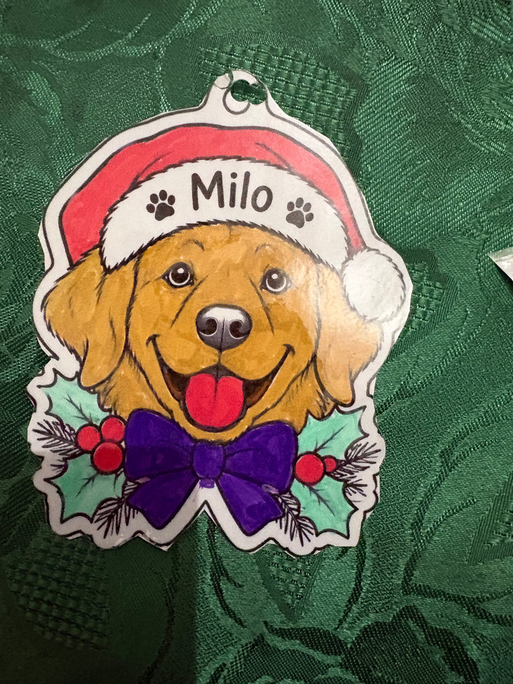 A pet Christmas ornament shaped like a Golden Retriever dog, featuring a red Santa hat with the name 'Milo' and paw prints, a purple bow tie, and holly accents, displayed on a textured green fabric background.