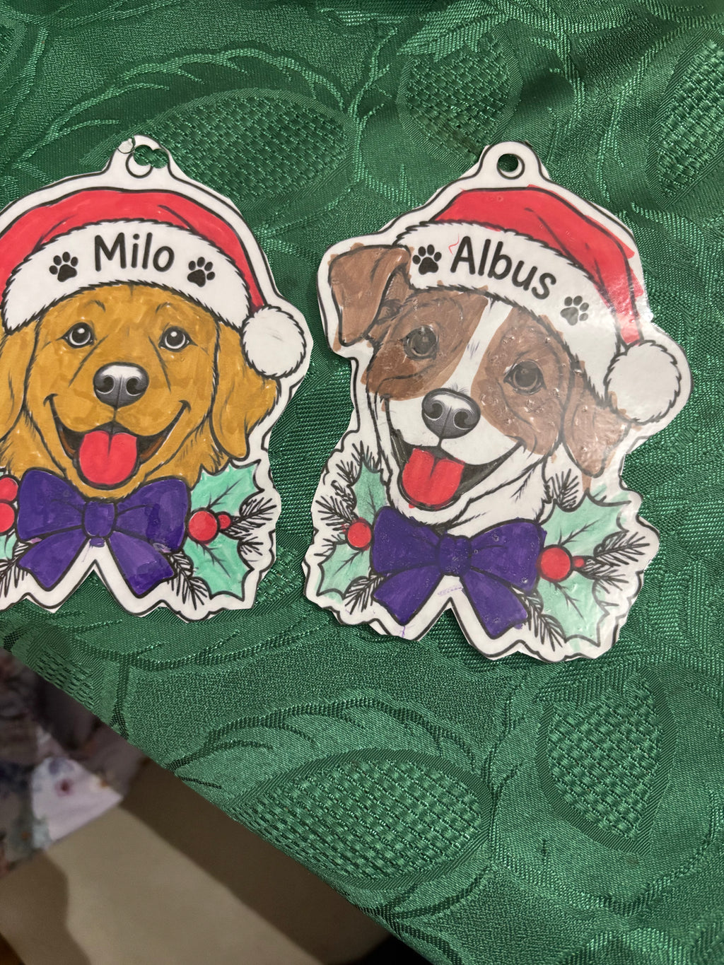 Two dog-shaped Christmas ornaments on a textured green fabric background. One ornament features a golden retriever dog wearing a Santa hat and purple bow tie, labeled 'Milo'.