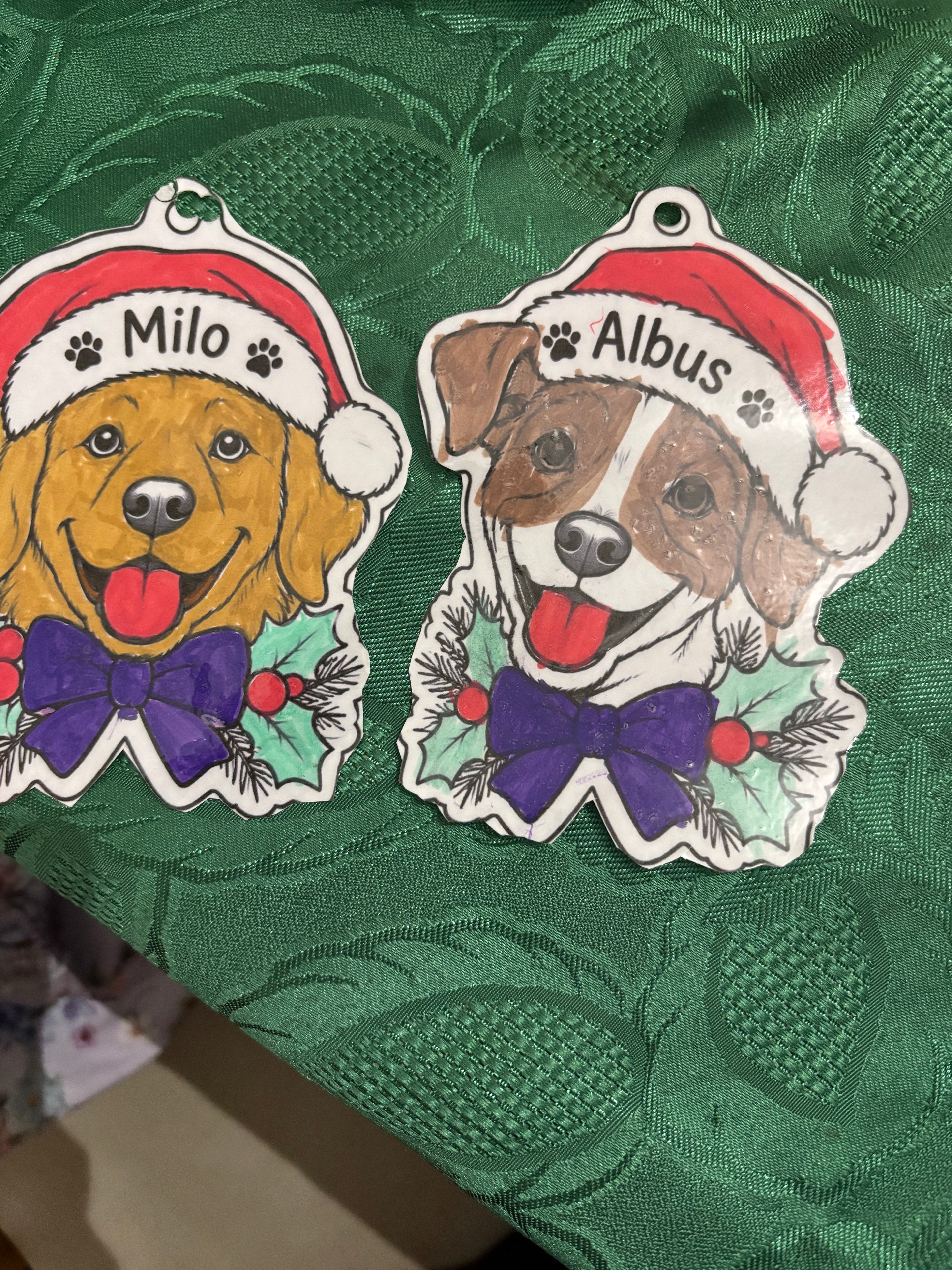 Two dog-shaped Christmas ornaments on a textured green fabric background. One ornament features a golden retriever dog wearing a Santa hat and purple bow tie, labeled 'Milo'.