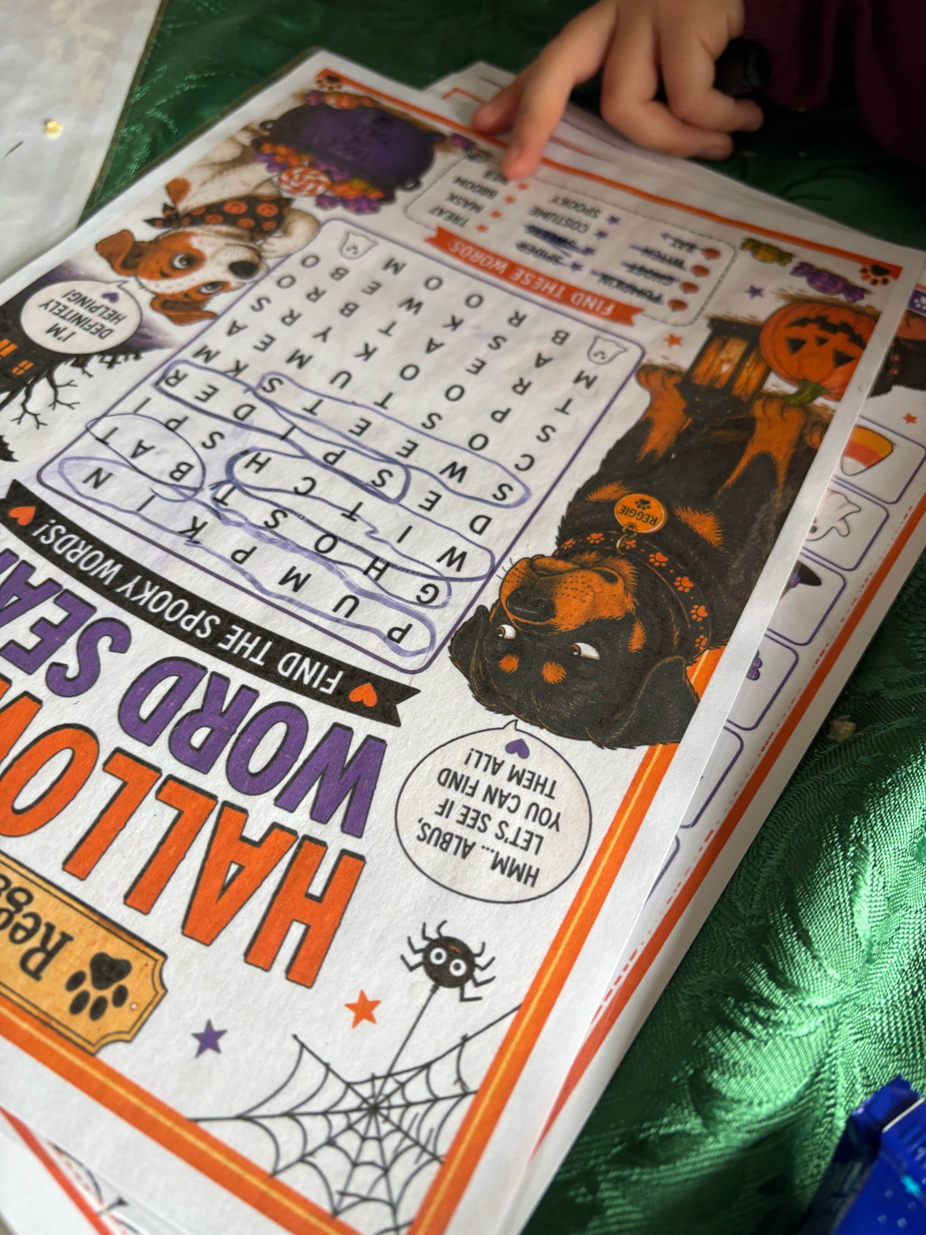 A close-up of a printed Halloween word search activity sheet being held by a child's hand over a shiny green surface. The page features spooky illustrations including a witch, pumpkins, a spider, and large Halloween-themed text.