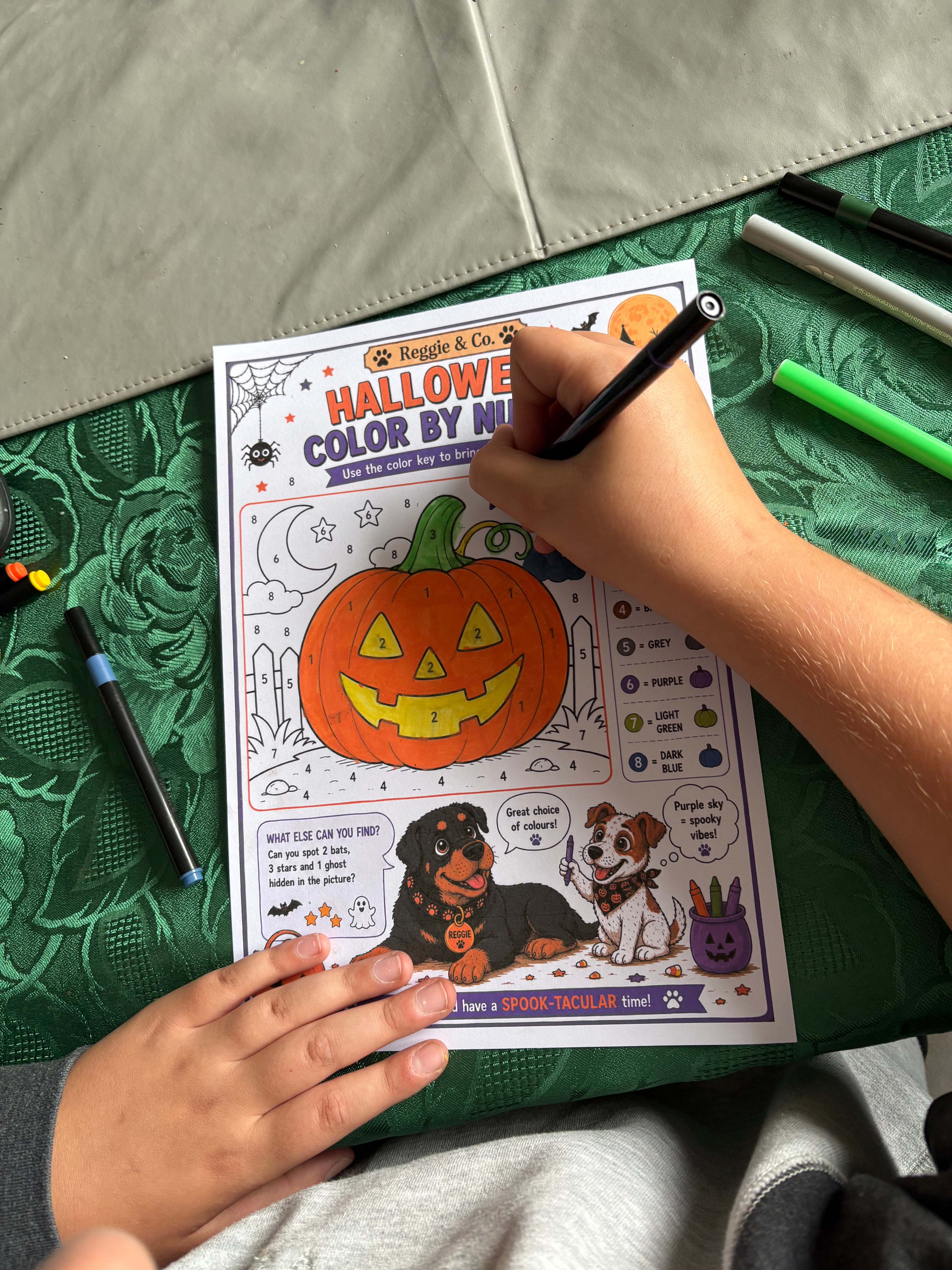 A person's hands are shown coloring a 'Halloween Color by Number' activity sheet with a black marker. The sheet features a jack-o'-lantern and cartoon dogs.
